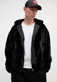 Black faux fur zip-up jacket with a hood, featuring a smooth quilted interior and side pockets, worn over a white shirt and black pants.