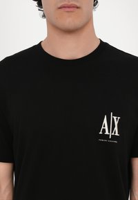 Svart bomull t-skjorte med rund hals og korte ermer, med en hvit brodert logo på venstre bryst. Myk tekstur.