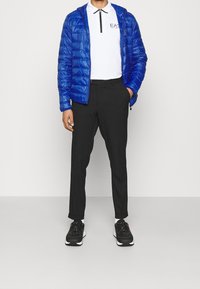 Blauer Pufferjacke, weißes Polo mit schwarzen Akzenten, schwarze Hose und schwarze Turnschuhe mit weißen Sohlen. Minimalistisches Design, taillierte Passform.