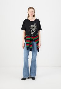 Juodas grafikos t-shirt su metaliniu spaudu, derinamas su šviesiai mėlynomis platėjančiomis džinsomis. Apie liemenį surišta raudona, žalia ir tamsiai mėlyna juostuota megzta palaidinė.