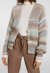 Sweter kardigan w beżowe i pastelowo-niebieskie paski, o luźnym kroju, szerokich rękawach, teksturowanej tkaninie i drewnianych guzikach. Połączony z brązowymi spodniami.