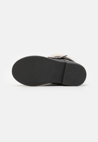 Semelle en caoutchouc noir avec une texture antidérapante. La tige est composée d'un matériau doux et clair avec des accents décoratifs. Design plat pour un confort optimal.