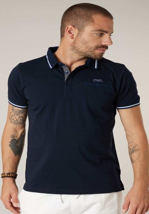 Homme aux cheveux courts et tatouages portant un polo bleu marine avec un liseré blanc et un pantalon blanc, regardant vers sa droite sur un fond uni.