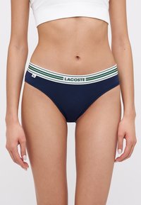 Tamsiai mėlyna medvilninė bikini maudymosi kostiumėlis su balta juostele, dekoruota žaliomis juostelėmis ir "LACOSTE" logotipu. Lygi tekstūra, klasikė fasonas.