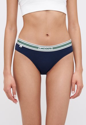 Marineblauwe katoenen bikini met een witte tailleband met groene strepen en het "LACOSTE" logo. Glad oppervlak, klassieke snit.
