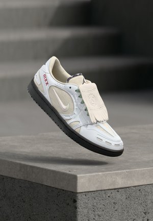 Λευκό και μπεζ αθλητικό παπούτσι Nike με μαύρη σόλα και λεπτομέρεια φραγμών που αιωρείται πάνω από μια τσιμεντένια άκρη με θολές σκάλες στο φόντο.