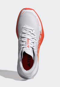 Scarpa atletica bianca leggera con accenti rossi, tomaia in mesh, suola texturizzata e sistema di allacciatura. Presenta il marchio "TERREX" di lato.