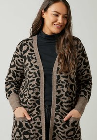 Valkoinen ja musta leopardi-kuvioinen neuletakki, avonainen etuosa, ribbikannet ja kaksi etutaskua. Valmistettu pehmeästä neulekankaasta.