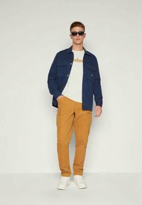 Camisa azul-marinho de botões com dois bolsos frontais, t-shirt branca com texto, calças cargo mostarda e sapatilhas brancas, usadas com óculos de sol.