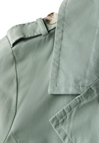 Jasnozielony materiał o gładkiej teksturze, z zaokrąglonym kołnierzykiem i dużym guzikiem. Szczegóły obejmują staranne szwy i dopasowany krój.