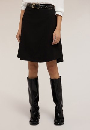 Vrouw draagt een zwarte kokerrok tot op de knie met een zwart, met studs versierd riem, een wit overhemd en glanzende zwarte laarzen tot aan de knie, staand tegen een effen achtergrond.