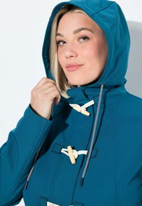 Kvinna med blont hår som bär en blå jacka med huva och vita togglar, håller i huvarets dragsko nära sitt ansikte.