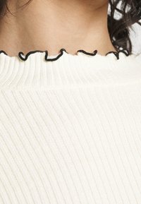 Närbild av en ribbad vit tröja med svart volangkant som bärs av en person med mörkt lockigt hår.