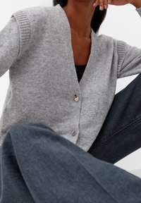 Grå ullkofta med djup V-hals, ribbade ärmar och två knappar i silverfärg. Tillsammans med mörka jeans, som har en mjuk textur.