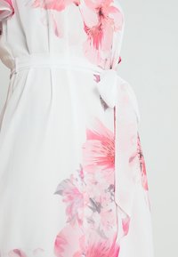 Robe blanche avec de grands motifs floraux roses et une ceinture nouée à la taille sur un fond clair.