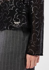 Διάφανη μαύρη μπλούζα με σπειροειδή ασημένια σχέδια, διαθέτοντας ετικέτα λογοτύπου. Συνδυάζεται με γκρι ριγέ παντελόνι, τονίζοντας τις υφασματικές αντιθέσεις.