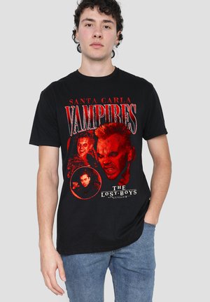 Young man wearing black T-shirt with red graphic of vampire faces and text "Santa Carla Vampires" and "The Lost Boys."