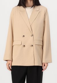 Femme portant un blazer beige croisé sur un pull côtelé crème et un pantalon noir, debout devant un fond blanc uni.