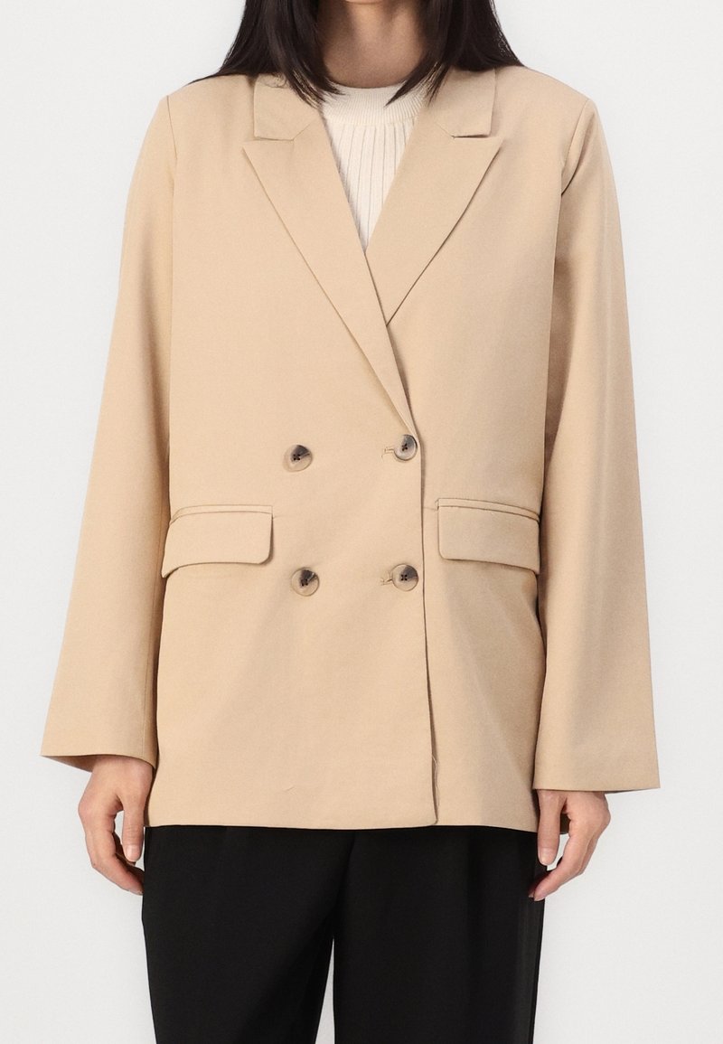 Femme portant un blazer beige croisé sur un pull côtelé crème et un pantalon noir, debout devant un fond blanc uni.