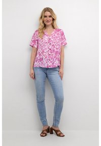 Blusa rosa a maniche corte con motivo floreale, bottoni sulla parte anteriore, abbinata a jeans azzurri chiari e sandali con tacco marroni.