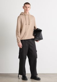 Hanorac bej cu șnururi, buzunar tip cangur și logo. Pantaloni cargo negri cu buzunare laterale și mansete reglabile. Geantă tote neagră ținută în mână.