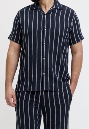 Homme portant une chemise bleu marine à manches courtes avec boutons et un pantalon assorti à rayures verticales blanches, posant avec une main dans la poche.