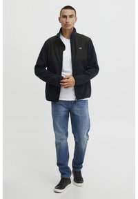 Giacca in pile navy con pannelli neri, colletto alto e chiusura a zip. Indossata con una t-shirt bianca e jeans blu, e sneaker nere.