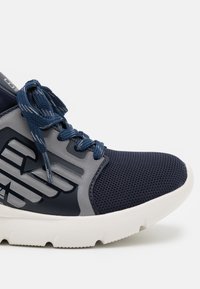 Marinblå atletiska skor med en ovandel i mesh, grå detaljer, texturerad yta, rund tå och vit gummisula. Har blå snören och logotypdetalj.