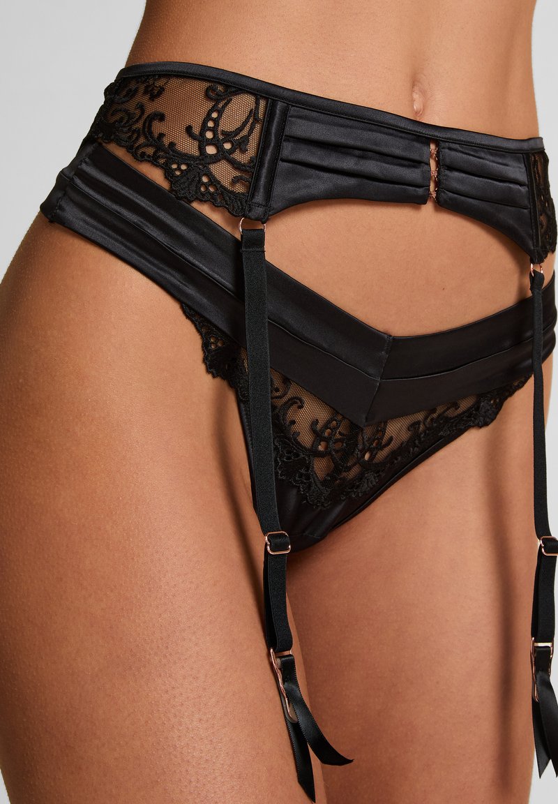 Zwart satijnen jarretel met doorschijnende meshpanelen en kantdetails. Voorzien van verticale plooien, verstelbare banden en roségouden metalen accenten.