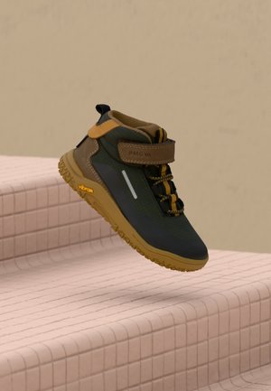 Chaussure de randonnée vert foncé et marron avec des accents jaunes et une semelle texturée, flottant au-dessus d'escaliers carrelés roses sur fond beige.