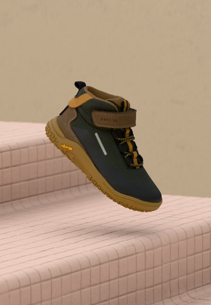 Chaussure de randonnée vert foncé et marron avec des accents jaunes et une semelle texturée, flottant au-dessus d'escaliers carrelés roses sur fond beige.