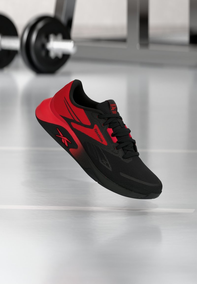 Μαύρο και κόκκινο αθλητικό παπούτσι Reebok με ελαφρύ σχεδιασμό, που αιωρείται πάνω από το πάτωμα ενός γυμναστηρίου με θολά βάρη στο φόντο.