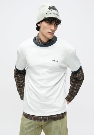 Fehér pamut póló tengerészkék gallérral, barna kockás ing fölött rétegezve. A modell kötött sapkát visel fekete grafikával. Texturált, laza megjelenés.