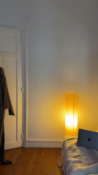 Hohe, zylindrische Lampe mit warmem gelbem Licht neben einer grauen Decke, einem weißen Sofa und einem Laptop auf einem Holzfußboden. Tür teilweise geöffnet.