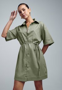 Oliivroheline lina segu särgimekko, millel on lühikesed varrukad, ees taskud, nööpkinnitus ja reguleeritava sobivusega seotud vöö.