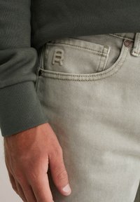 Mano descansando junto al bolsillo de unos jeans de mezclilla gris claro con un pequeño logo bordado y la manga de un sudadera de canalé verde oliva.