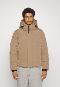 Giacca puffer stile berretto di colore marrone chiaro, con zip frontale, colletto alto e ampie sezioni trapuntate. Include cordini regolabili.