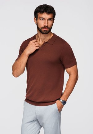 Homme aux cheveux bouclés et foncés avec une barbe, portant un polo marron à manches courtes et un pantalon gris clair, posant avec une main dans la poche et l'autre près du col.