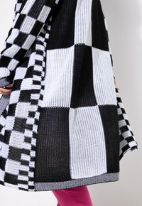 Cardigan tricoté avec un motif à damier noir et blanc, featuring des rayures contrastantes et une coupe décontractée, associé à un pantalon rose.