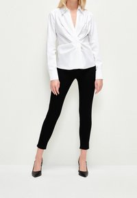 Chemise blanche à manches longues avec un design torsadé à l'avant, associée à un pantalon noir ajusté et des talons noirs pointus. Texture de tissu lisse.