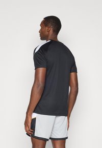 Μαύρη αθλητική μπλούζα με κοντά μανίκια και λευκή λεπτομέρεια στους ώμους, σε συνδυασμό με γκρι σορτς. Το υλικό φαίνεται ελαφρύ και αναπνέει.