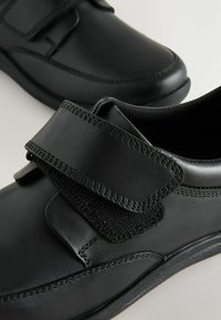 Chaussures noires en matériau synthétique avec une finition lisse, dotées d'une sangle Velcro et de coutures détaillées le long des bords.