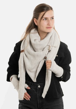 Chunky gebreide off-white sjaal met een textuurpatroon, losjes gedrapeerd. Geaccentueerd met een houten toggle en een grijze knop. Draagt met een zwarte jas.