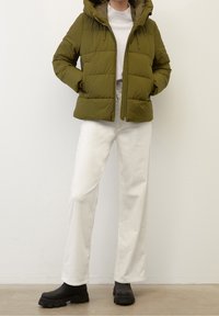 Olivgrön vadderad jacka med huva, framdragkedja och elastiska dragskor. Kombinerad med vita byxor med vida ben och svarta klumpiga stövlar.