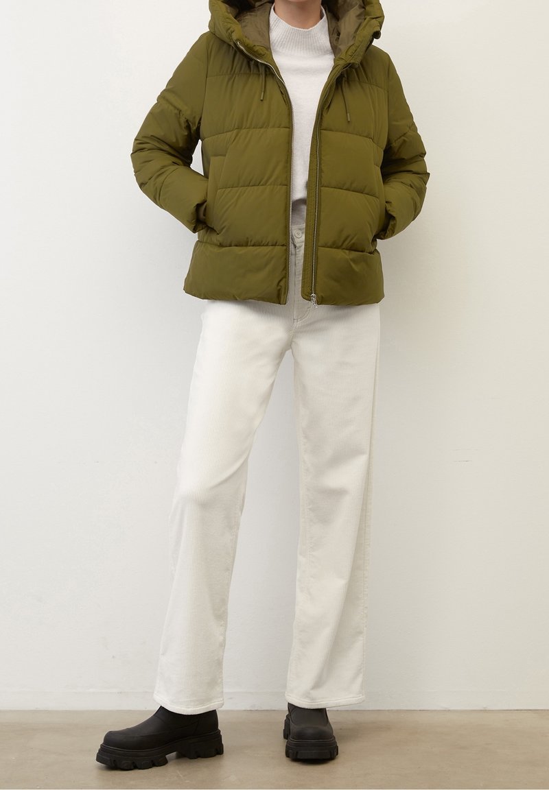 Olivgrön vadderad jacka med huva, framdragkedja och elastiska dragskor. Kombinerad med vita byxor med vida ben och svarta klumpiga stövlar.