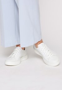Sneakers bianchi in pelle con finitura liscia, caratterizzati da una punta rotonda, lacci bianchi piatti e una suola spessa e testurizzata. Design lucido e accenti minimi.