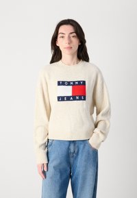 Ecru dzianinowy sweter z ryflem na rękawach i okrągłym dekoltem. Centralne logo w czerwono-białym-nawigacyjnym wzorze. Połączony z jasnoniebieskimi dżinsami.