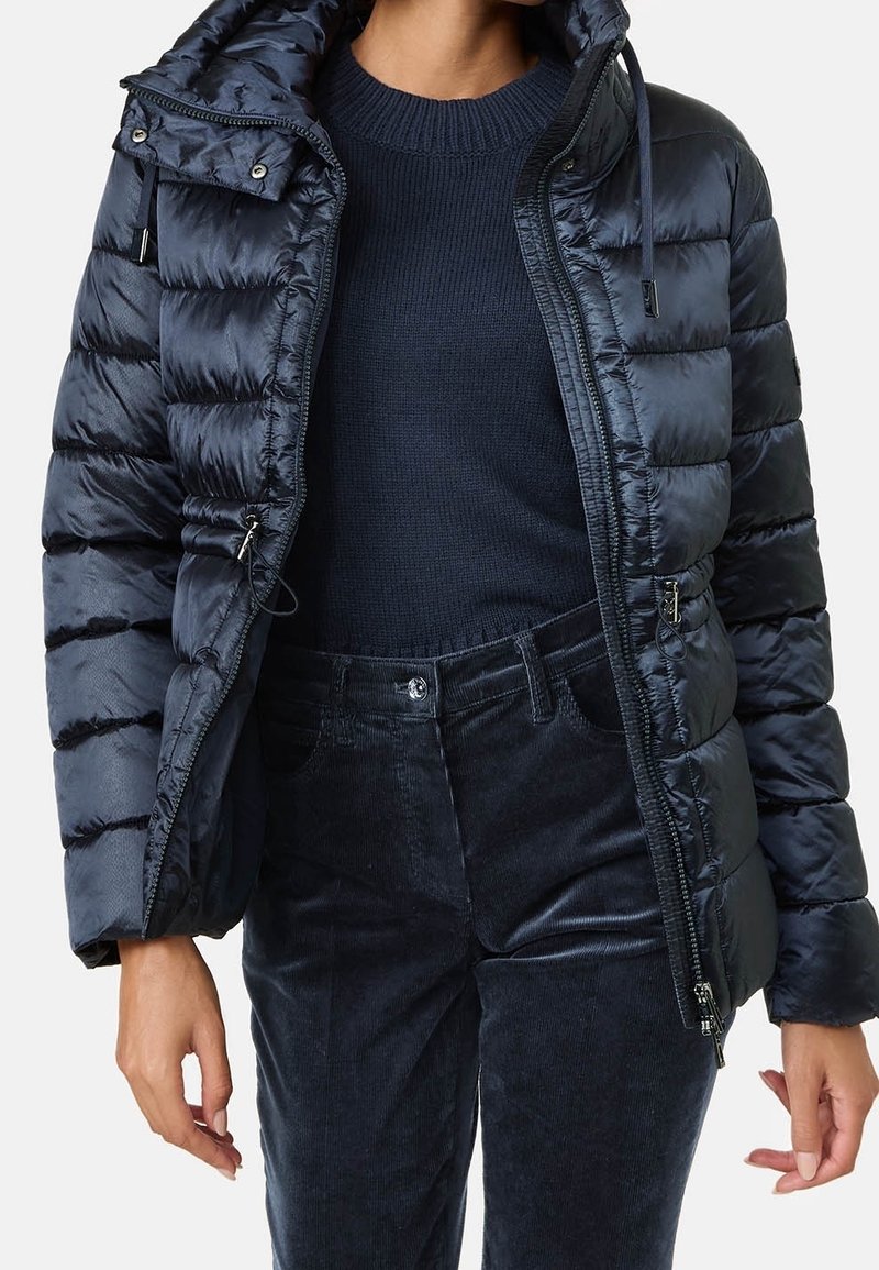 Chaqueta acolchada azul marino con capucha, que presenta un acabado brillante, patrón de acolchado, cierre frontal con cremallera y dobladillo ajustable. Combinada con un suéter azul marino y pantalones.