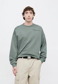 Hombre con pantalones cargo beige, sudadera verde con pequeño texto en el pecho, cinturón negro y gorra beige puesta al revés, de pie contra un fondo blanco.