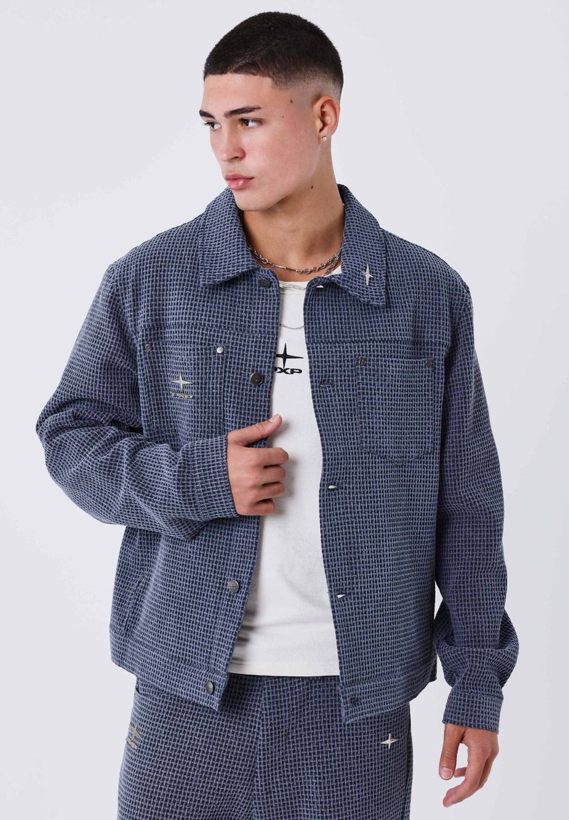 Giacca a quadri blu navy in tessuto strutturato con chiusura a bottoni, due tasche frontali e vestibilità rilassata; indossata sopra una t-shirt chiara.